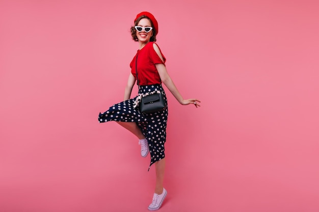 Portrait en pied d'un modèle féminin de bonne humeur en béret rouge français dansant avec le sourire Incroyable fille bouclée en lunettes noires sautant en studio