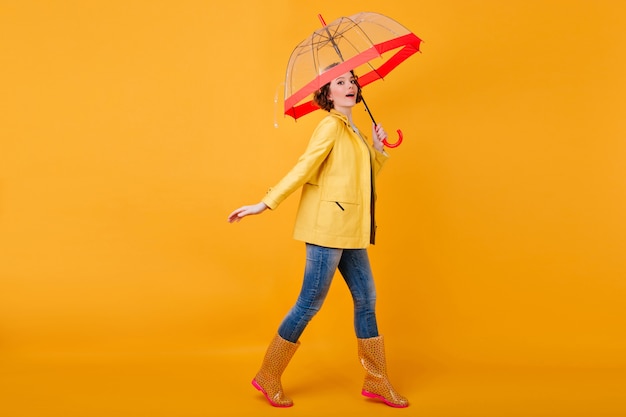 Portrait en pied d'un modèle féminin caucasien en veste jaune et chaussures en caoutchouc. Photo de Studio d'une fille insouciante aux cheveux ondulés dansant avec un parapluie.