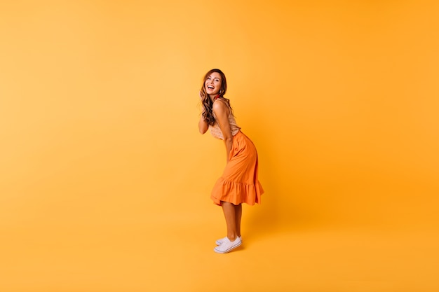 Portrait en pied d'une superbe fille bouclée en jupe orange. Portrait en studio de dame positive dansant sur jaune.