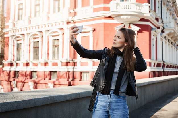 Portrait en plein air de belle femme debout dans le centre-ville, posant tout en tenant le smartphone et en prenant selfie, portant des vêtements à la mode