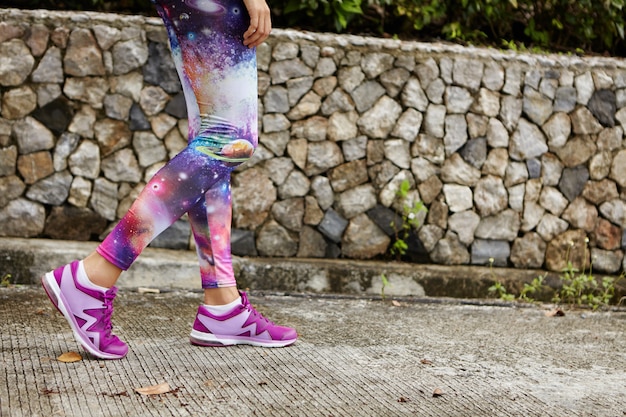 Portrait en plein air de coureuse avec des jambes athlétiques en forme portant des baskets violettes marchant le long d'un chemin en béton dans un parc urbain, reprenant son souffle après un entraînement intensif, se préparant pour le marathon