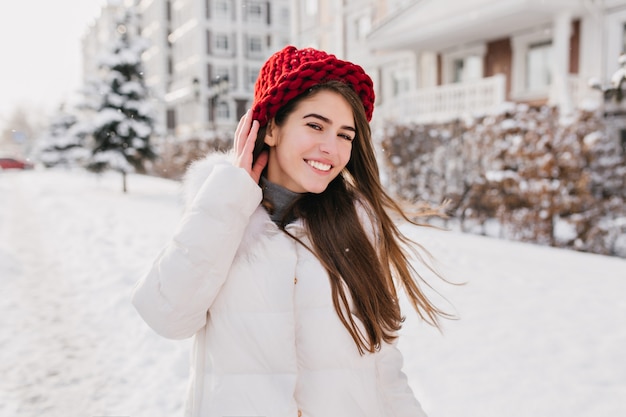 Portrait en plein air de dame aux cheveux longs heureux en bonnet rouge marchant dans la rue en week-end enneigé. Photo de femme mignonne en riant en manteau d'hiver blanc s'amusant dans la froide matinée.