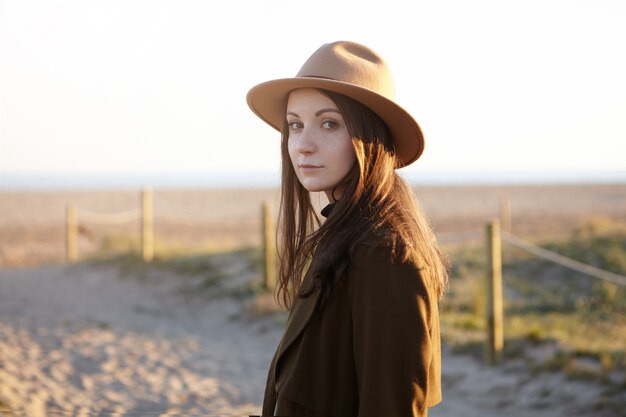 Portrait en plein air de l'élégante jeune femme européenne portant un chapeau à la mode et un manteau noir à la recherche d'un sourire subtil tout en ayant une belle soirée à pied au bord de la mer, rêvant et admirant le coucher du soleil