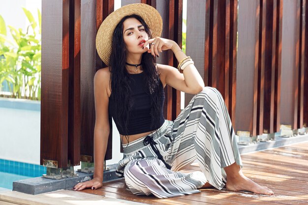 Portrait en plein air d'été d'une femme sensuelle en chapeau de paille, élégant pantalon rayé posant dans une villa de luxe près de la piscine