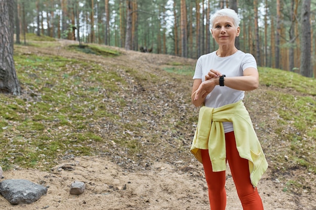 Portrait en plein air de femme d'âge moyen active confiante en vêtements de sport à l'aide d'une montre intelligente surveillance du pouls ou de la fréquence cardiaque lors de l'exercice dans le parc.