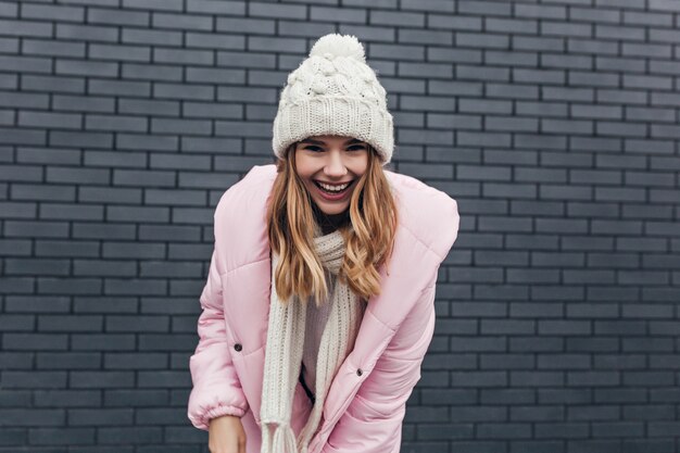 Portrait en plein air de femme blithesome en manteau rose. Jolie fille blonde au chapeau d'hiver posant devant le mur de briques.