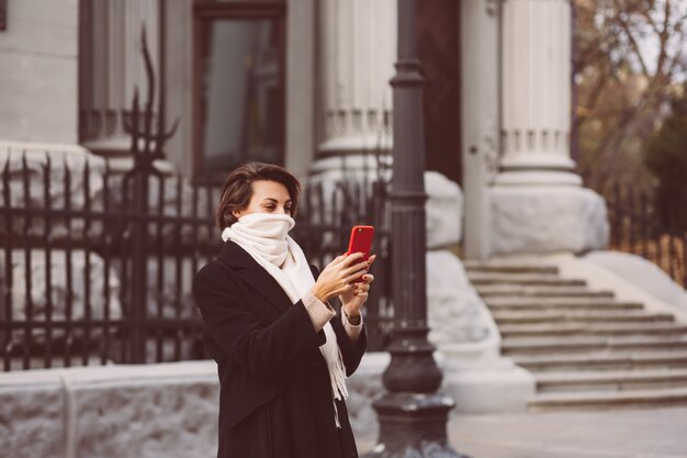 Portrait en plein air de femme en manteau d'hiver noir et écharpe blanche dans la rue, tenant un téléphone mobile.