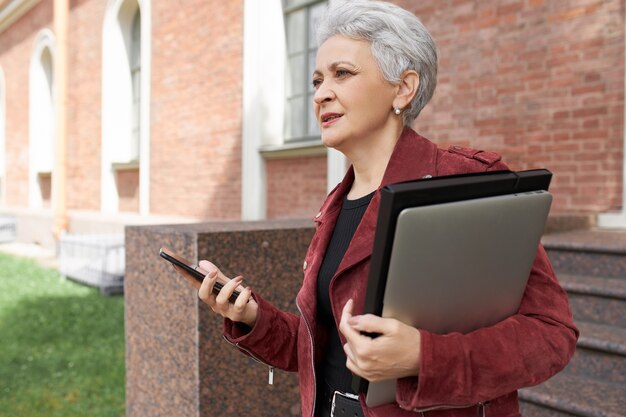 Portrait en plein air de femme mûre sérieuse gestionnaire étant pressé de bureau, posant à l'extérieur avec un ordinateur portable, commande de taxi en ligne via l'application sur téléphone mobile