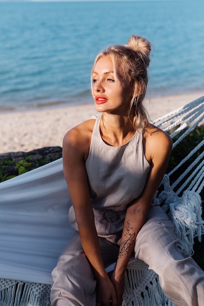 Portrait en plein air de femme de race blanche en combinaison classique avec rouge à lèvres par hamac en vacances à l'extérieur de l'hôtel Villa, côté mer