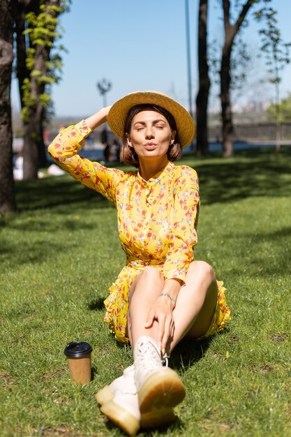 Portrait en plein air de femme en robe d'été jaune et chapeau assis sur l'herbe dans le parc