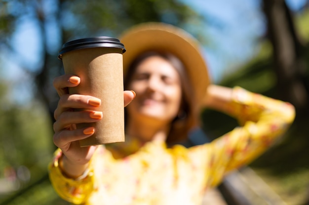 Portrait en plein air de femme en robe d'été jaune et chapeau avec tasse de café dans le parc