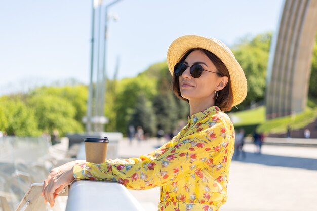 Portrait en plein air de femme en robe d'été jaune et chapeau avec tasse de café en profitant du soleil, se dresse sur le pont avec vue imprenable sur la ville