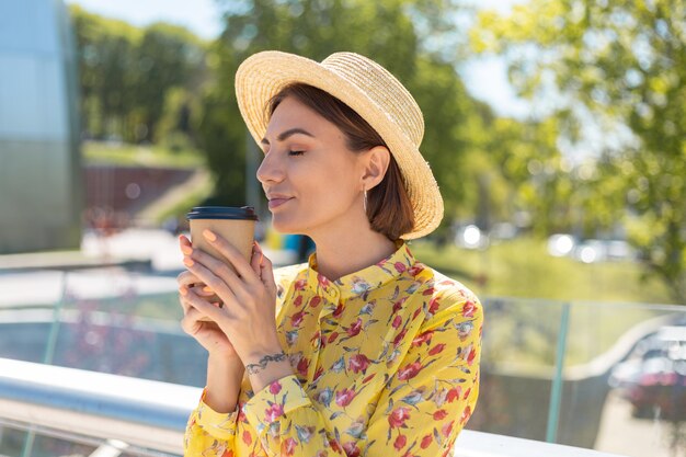 Portrait en plein air de femme en robe d'été jaune et chapeau avec tasse de café en profitant du soleil