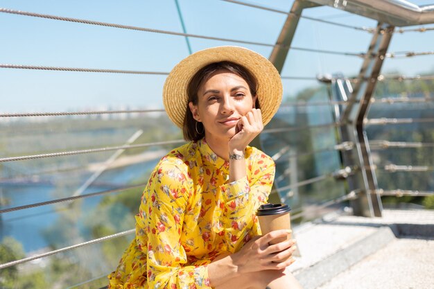 Portrait en plein air de femme en robe d'été jaune et chapeau avec tasse de café en profitant du soleil