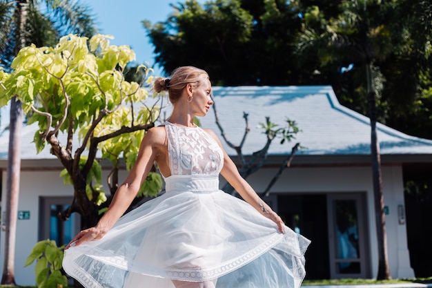 Portrait en plein air de femme en robe de mariée blanche à la villa en journée ensoleillée, vue tropicale