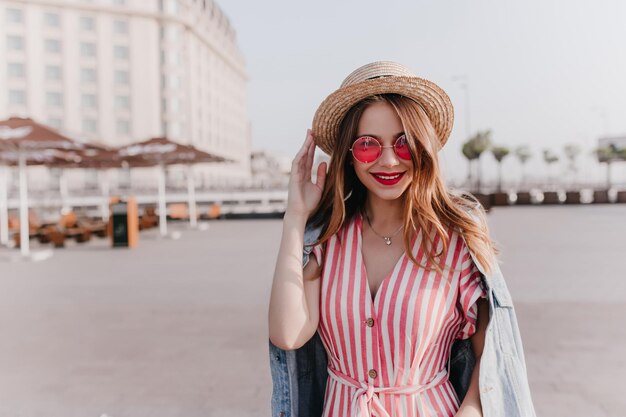 Portrait en plein air d'une fille élégante et agréable au chapeau vintage debout sur fond de ville Modèle féminin de bonne humeur dans des lunettes de soleil roses profitant d'une chaude journée de printemps