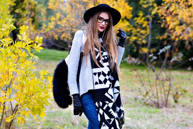 Portrait en plein air d'une fille excitée porte à la mode à larges bords et debout dans une pose confiante. Jolie jeune femme à lunettes posant sur fond de nature automne.