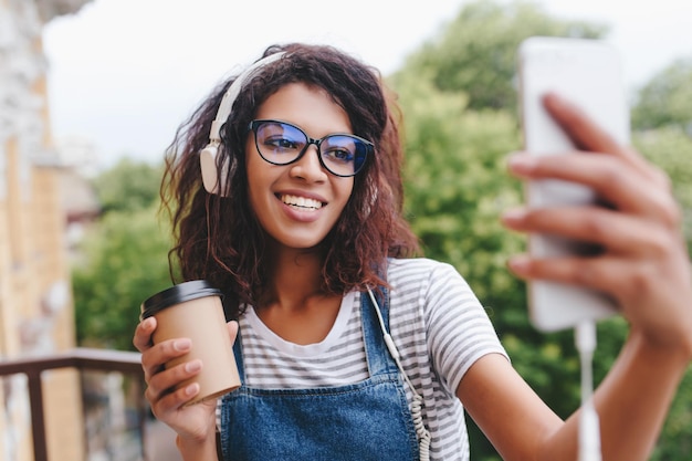 Photo gratuite portrait en plein air d'une fille joyeuse à la peau brune passant du temps à l'extérieur et se prenant en photo avec des arbres en arrière-plan. jolie jeune femme avec une tasse de café faisant un selfie tout en écoutant de la musique.