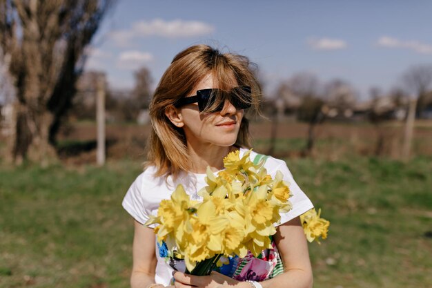 Portrait en plein air d'une fille souriante à lunettes de soleil tient des fleurs jaunes regarde loin et souriant