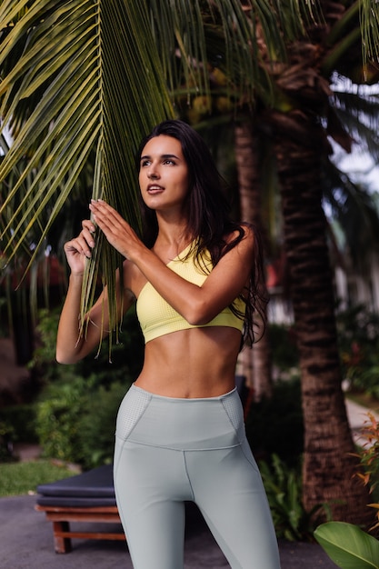 Portrait En Plein Air De Fit Slim Sportive Bronzée Jolie Femme De Race Blanche Dans Le Sport Haut Jaune Et Leggings Sur Palmier