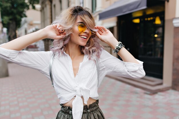 Portrait en plein air d'une jeune femme gaie en chemise blanche vintage et lunettes jaunes portant un élégant sac à dos en argent. Fille blonde assez bouclée dans de jolis bijoux passant du temps à l'extérieur à marcher près du café.