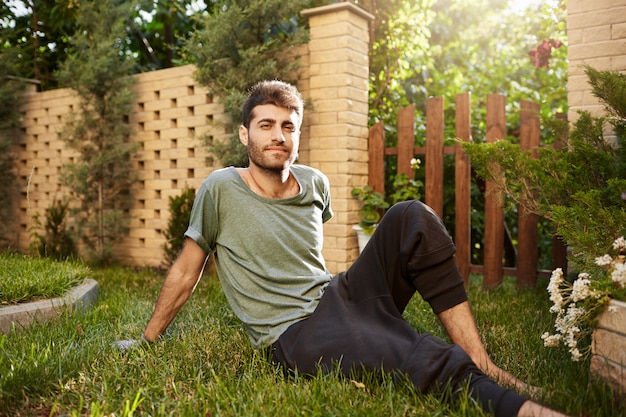 Portrait en plein air de jeune jardinier mâle caucasien barbu attrayant souriant, assis sur l'herbe dans le jardin.