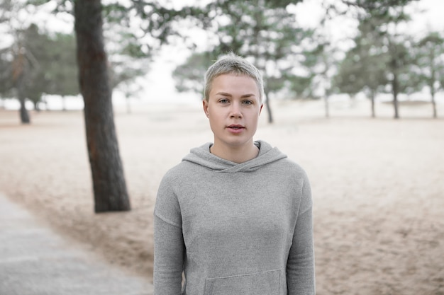 Portrait en plein air de jolie femme blonde aux cheveux courts à la mode portant un sweat à capuche gris à la mode reprenant son souffle pendant le repos tout en courant dans le parc seul, travaillant sur l'endurance et la force