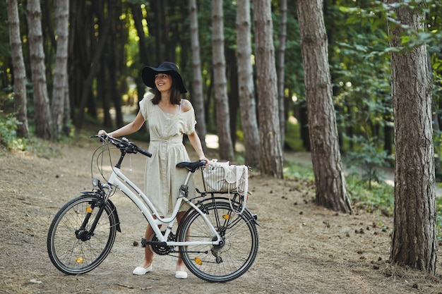 Portrait en plein air de séduisante jeune brune au chapeau sur un vélo.