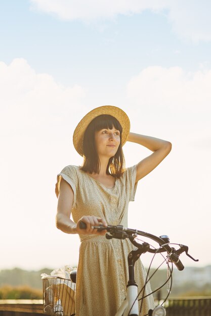 Portrait en plein air de séduisante jeune brune au chapeau sur un vélo.