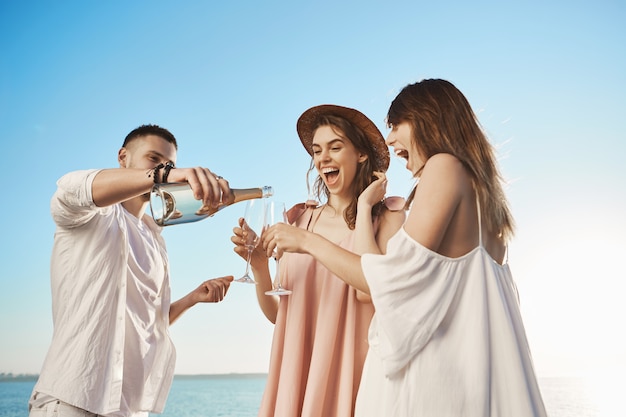 Portrait en plein air de trois jeunes adultes, buvant du champagne et souriant largement tout en se reposant au bord de la mer. Beau barbu poors boit à ses amis des verres, applaudissant à leur avenir heureux