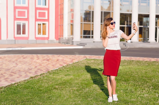 Portrait pleine hauteur d'une jeune fille souriante debout tenant un smartphone et faisant un selfie. La fille porte un t-shirt blanc, une jupe rouge et des lunettes de soleil foncées. La fille est dans le centre-ville.