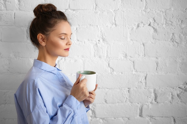 Portrait de profil d'élégante jeune femme d'affaires fatiguée avec maquillage et chignon relaxant au bureau avec une tasse de boisson fraîche pendant la pause-café, fermant les yeux, posant au mur de briques, tenant une tasse