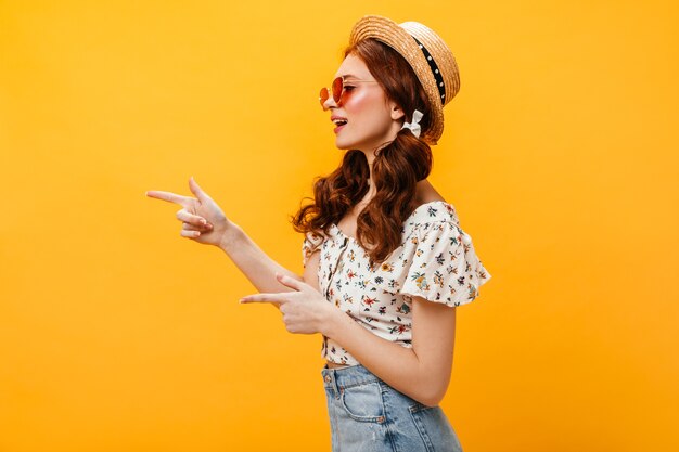 Portrait de profil de femme au chapeau de paille et lunettes. Dame avec deux queues de cheval pointant les doigts vers la gauche.