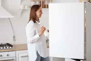 Photo gratuite portrait de profil d'une jolie femme aux cheveux noirs vêtue d'une chemise blanche, regardant souriant à l'intérieur du réfrigérateur avec des émotions positives, tenant une assiette dans les mains, posant avec un ensemble de cuisine en arrière-plan.