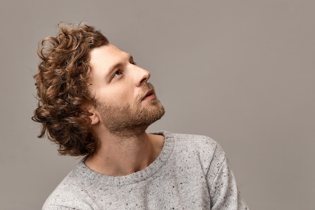 Portrait de profil de magnifique beau jeune homme avec des traits parfaits, des poils et des cheveux rougeâtres posant au mur de fond blanc en cavalier, regardant avec une expression faciale rêveuse