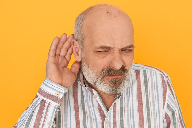 Portrait d'un retraité masculin barbu frustré fronçant en chemise rayée gardant la main à son oreille, écoutant attentivement, essayant d'entendre parler indistinct. Problèmes d'audition et écoute clandestine