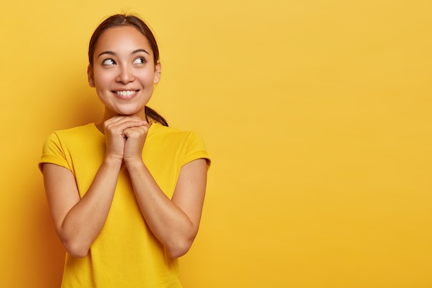 Photo gratuite portrait de rêve fille ethnique satisfaite concentrée de côté avec un sourire heureux, tient les mains sous le menton, regarde avec une expression d'espoir, croit en mieux, porte un t-shirt jaune décontracté, se tient en