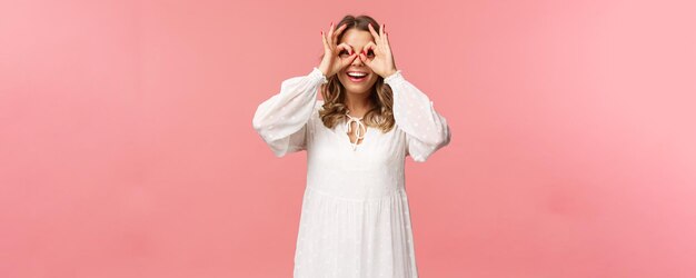 Portrait de rêveuse mignonne et drôle jeune fille blonde voyant quelque chose d'intéressant à la recherche de signes corrects comme faisant un masque de verre avec les doigts sur les yeux souriant fond rose amusé