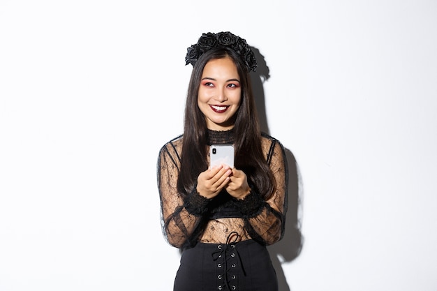 Portrait de la ruse belle femme asiatique, sorcière en robe de dentelle gothique à l'aide de téléphone mobile, souriant et regardant dans le coin supérieur gauche, debout sur un mur blanc