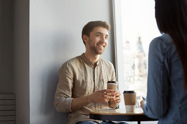 Portrait de séduisant jeune homme non rasé aux cheveux noirs, souriant, buvant du café et écoutant des histoires de petite amie sur une dure journée de travail. Mode de vie, concept de relation