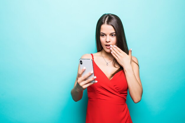 Portrait si une jeune fille en robe choquée à la recherche de téléphone mobile isolé sur mur bleu