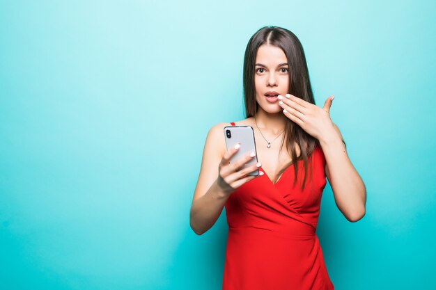 Portrait si une jeune fille en robe choquée à la recherche de téléphone mobile isolé sur mur bleu