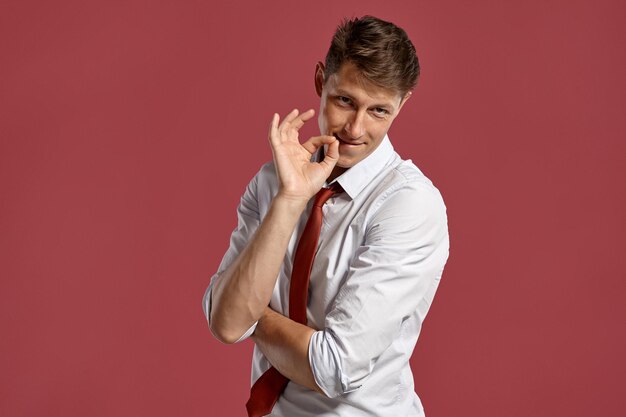 Portrait en studio d'un beau jeune homme dans une chemise blanche classique et une cravate rouge, souriant et montrant le signe ok tout en posant sur un fond rose. Coupe de cheveux élégante. Concept d'émotions sincères. Copiez l'espace.