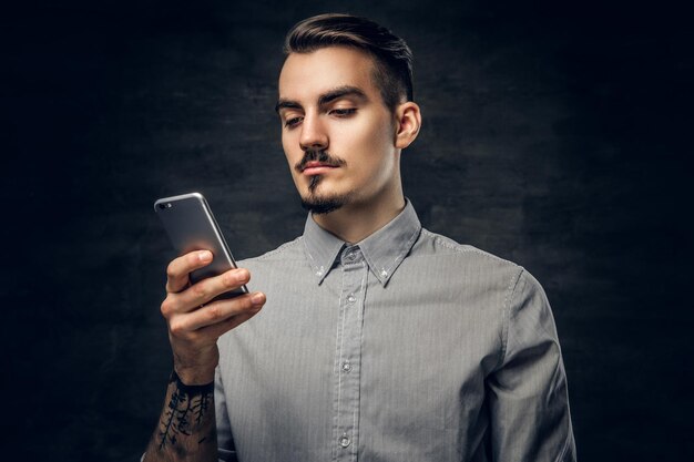 Portrait en studio d'un beau mâle hipster barbu avec un tatouage sur son bras à l'aide d'un smartphone.