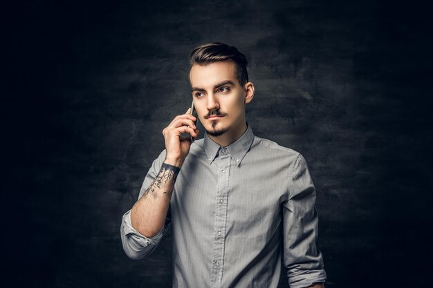 Portrait en studio d'un beau mâle hipster barbu avec un tatouage sur son bras parle sur un smartphone.