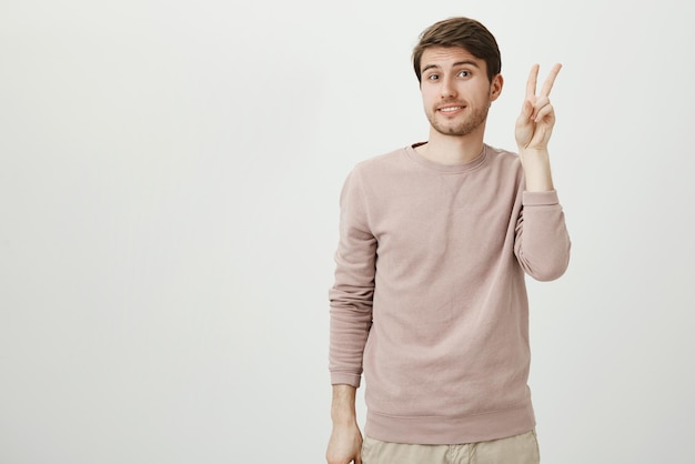 Portrait en studio d'un beau modèle masculin mignon montrant un geste de victoire ou de paix tout en souriant maladroitement