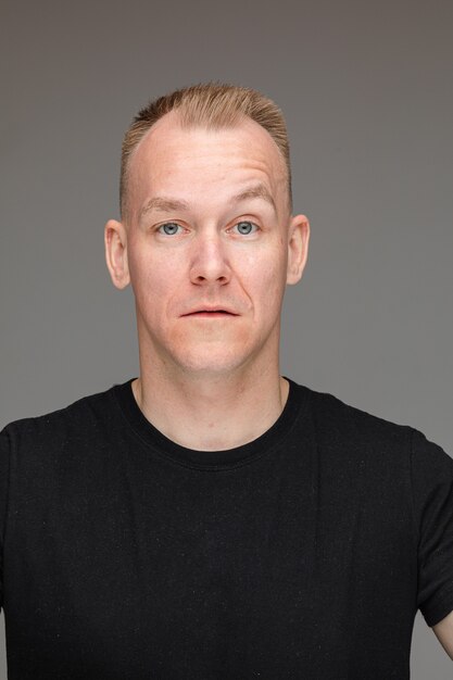 Portrait en studio d'un bel homme aux cheveux blonds courts et aux yeux bleus en t-shirt noir regardant la caméra avec les sourcils levés, montrant la confiance et l'arrogance.