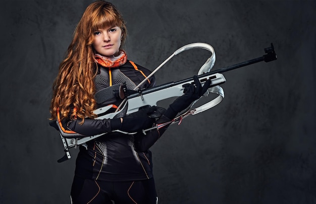 Portrait en studio d'une championne de biathlon à tête rouge tenant une arme à feu sur fond gris.