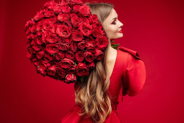 Portrait en studio sur l'épaule d'une magnifique jeune brune aux lèvres brillantes en robe rouge tenant un gros bouquet de roses rouges et souriant à la caméra sur fond rouge Isoler sur rouge