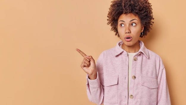 Photo gratuite le portrait en studio d'une femme aux cheveux bouclés choquée et impressionnée a surpris l'expression faciale pointe l'index sur l'espace de copie pour votre contenu publicitaire reste sans voix. omg regarde ça